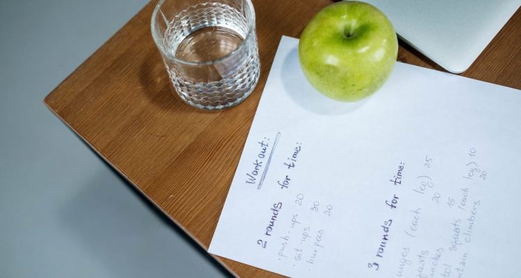 A green apple, glass of water, and workout plan on a wooden desk symbolize healthy living.